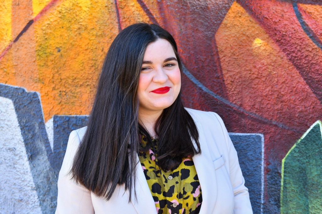 A woman with long dark hair smiles in front of a colourful mural, wearing a light blazer over a patterned shirt.